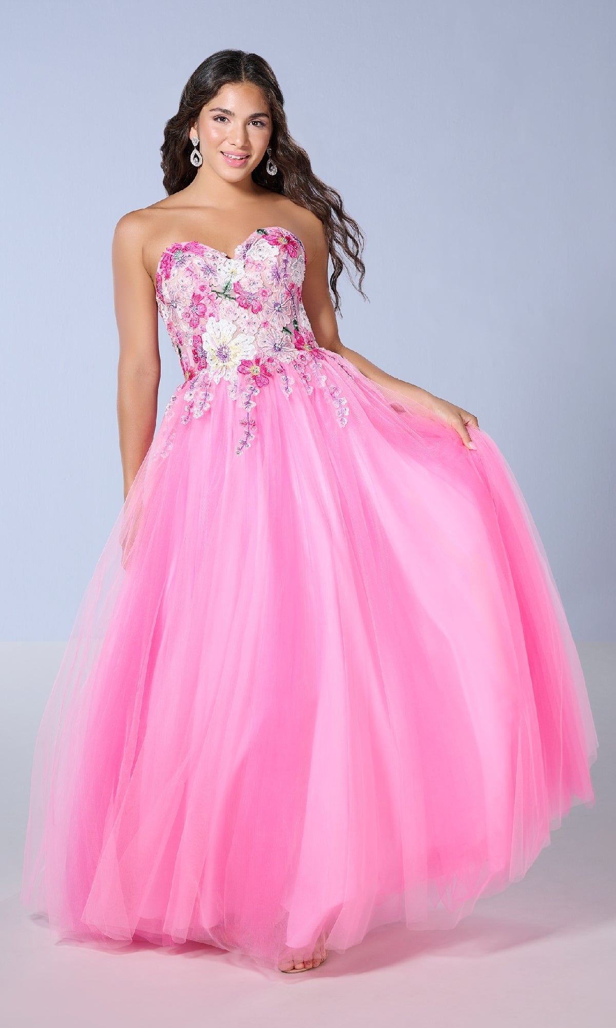 A woman in the Long Prom Dress: Tiffany 16142—featuring a strapless tulle skirt, floral lace embroidery, and a sheer corset bodice—stands against a plain light background, holding out her dress.